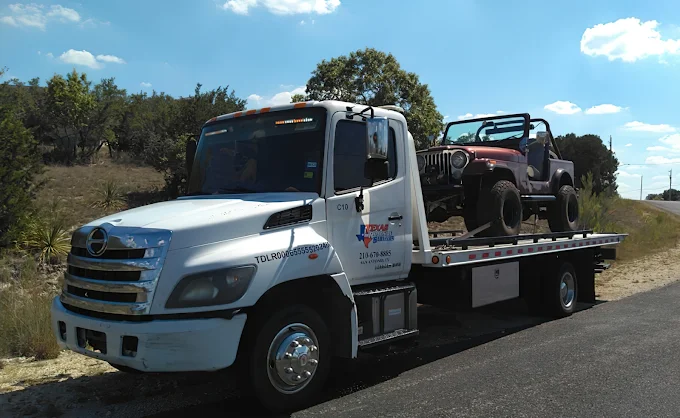 Jeep CJ loaded on a white flatbed tow truck on a Hill Country road